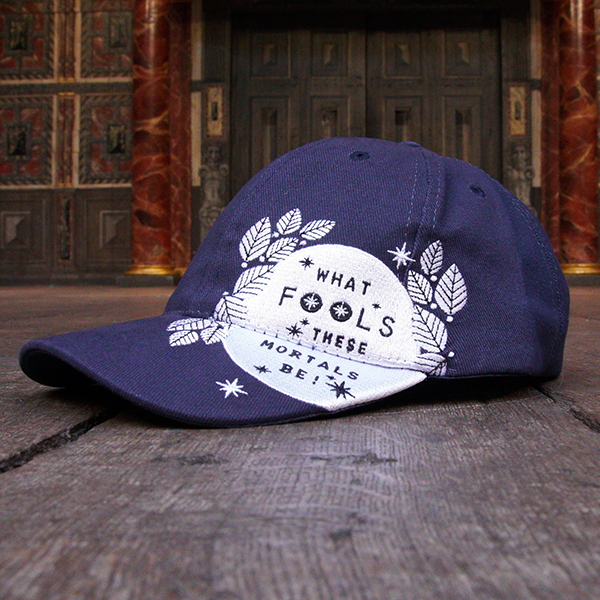 Navy blue six panel baseball cap with an embroidered motif in white across the left side of the cap and peak. The motif consists of the full moon surrounded by stylised leaves and stars. Across the face of the moon is a quote from Shakespeare play, A Midsummer Night's Dream, (what fools these mortals be!) in blue.