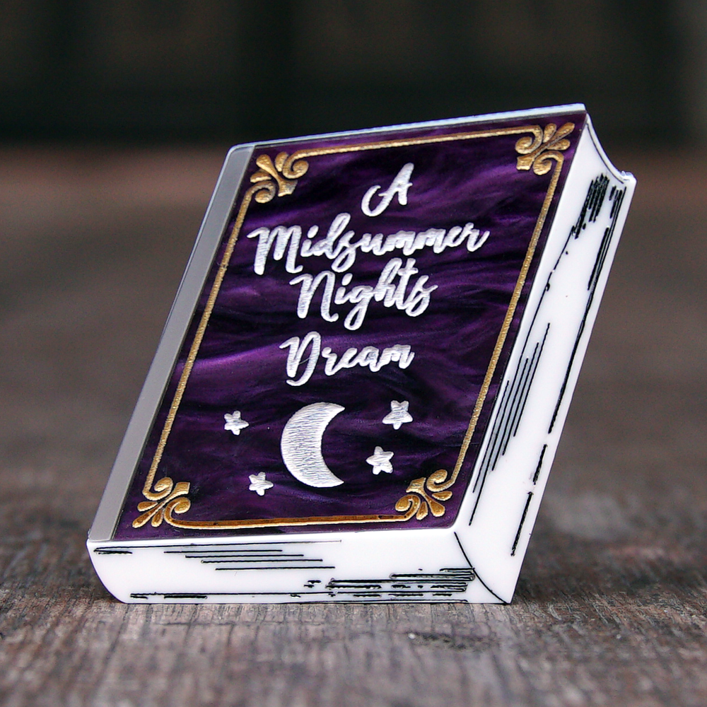 The brooch made of acrylic Perspex. The brooch is in the shape of a book so tat you can see the cover and the pages edges. The cover of the book is made of marbled purple Perspex and has a gold frame engraved around the edge. In the centre of the cover is 'A Midsummer Nights Dream' engraved in a white script. Below the title is engraved in white a crescent moon and four stars. The pages are made of white Perspex. 