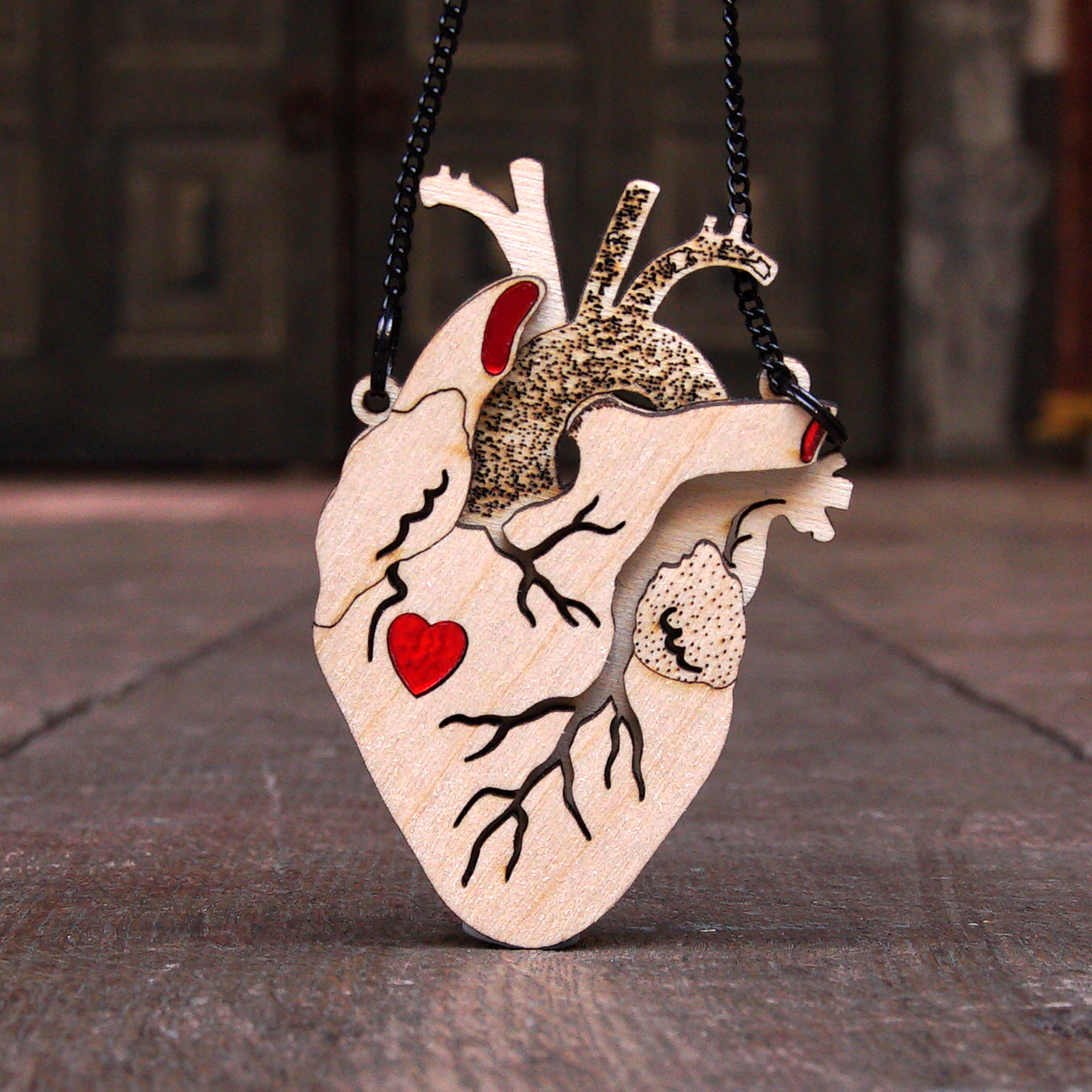 
                  
                    Pendant in the shape of an anatomical heart. The pendant is made of birch wood in several layers, giving the appearance of three dimensions. The top layer has channels cut to represent veins and the under layers are textured. A cutout in the shape of a graphic heart sits about halfway down the pendant and is filled in with bright red Perspex. the pendant is on a black chain.
                  
                