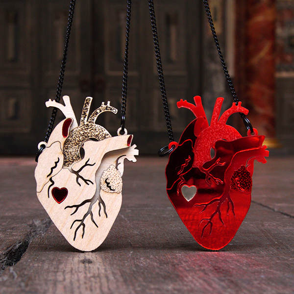 Two Pendants in the shape of an anatomical heart. One pendant is made of birch wood in several layers, giving the appearance of three dimensions. The top layer has channels cut to represent veins and the under layers are textured. A cutout in the shape of a graphic heart sits about halfway down the pendant and is filled in with bright red Persex. The second pendant is made in a similar way using bright red Perspex with a silver Perspex graphic heart cutout. Both pendants  are on black chains.