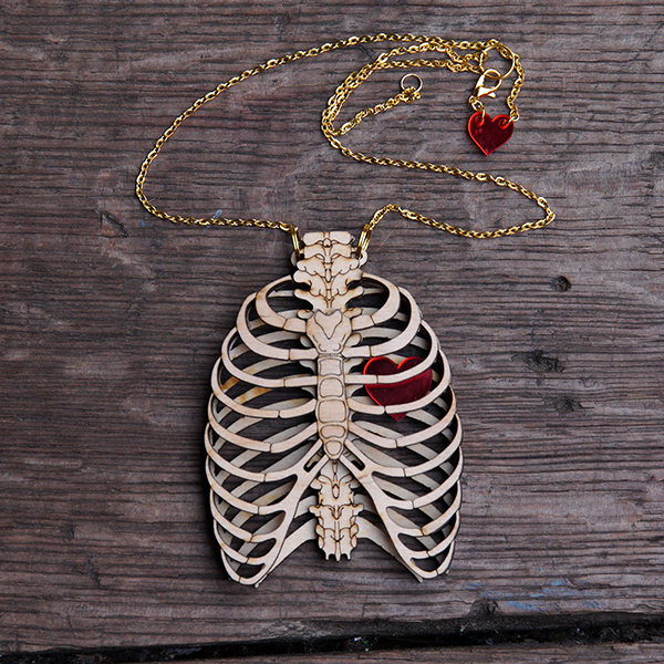 An anatomical human ribcage pendant made of birch wood. The pendant is made from two layers of laser-cut wood so that it looks as though the rib cage is 3D and you can see the back ribs and spine through the laser-cut ribs at the front. The wood is engraved to add detail to the bones including on the spine and breast bone. A red Perspex heart sits between the two layers of ribs. The pendant is hung on a gold coloured chain.