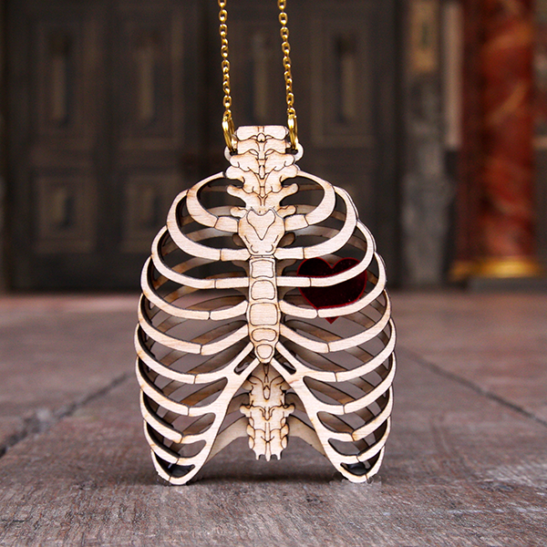An anatomical human ribcage pendant made of birch wood. The pendant is made from two layers of laser-cut wood so that it looks as though the rib cage is 3D and you can see the back ribs and spine through the laser-cut ribs at the front. The wood is engraved to add detail to the bones including on the spine and breast bone. A red Perspex heart sits between the two layers of ribs. The pendant is hung on a gold coloured chain.