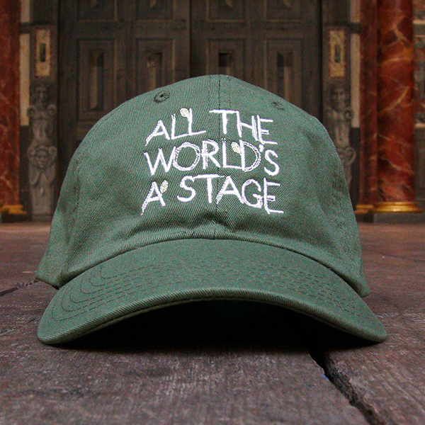 Khaki green six-panel baseball cap with a pre-curved peak. Embroidered on the front of the cap is a quote from Shakespeare play, As You Like It (all the world's a stage) in white hand-drawn capital letters.