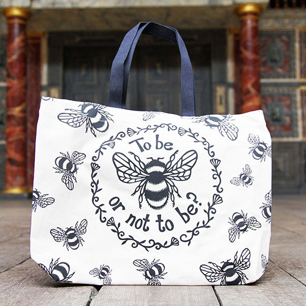 Tote bag made of heavyweight unbleached cotton canvas with mid-length black webbing handles. The bag is printed with a black design adapted from an original print of a bumble bee with a striped body and paneled wings, and a quote from Shakespeare play, Hamlet (To be or not to be?). In a ring around the bee and the quote are flowers and leaves. Outside this main design are a number of smaller bees flying in different directions.