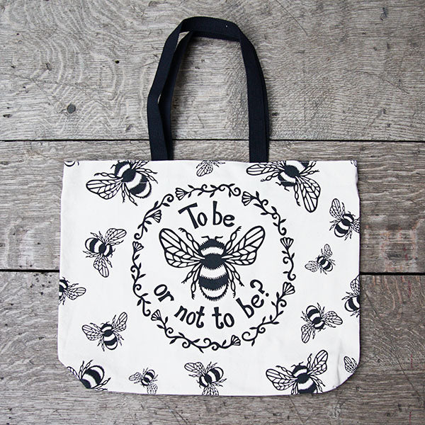Tote bag made of heavyweight unbleached cotton canvas with mid-length black webbing handles. The bag is printed with a black design adapted from an original print of a bumble bee with a striped body and paneled wings, and a quote from Shakespeare play, Hamlet (To be or not to be?). In a ring around the bee and the quote are flowers and leaves. Outside this main design are a number of smaller bees flying in different directions.