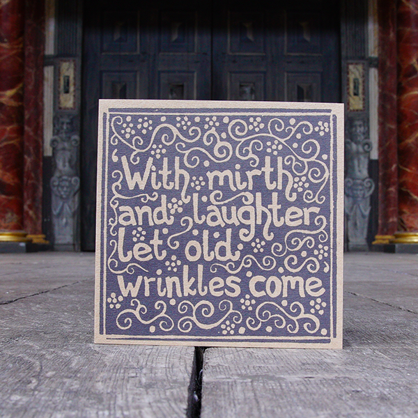 Kraft card greeting card with a lino print in black of a quote from Shakespeare play, The Merchant of Venice (With mirth and laughter let old wrinkles come). The quote is surrounded by vines and flowers