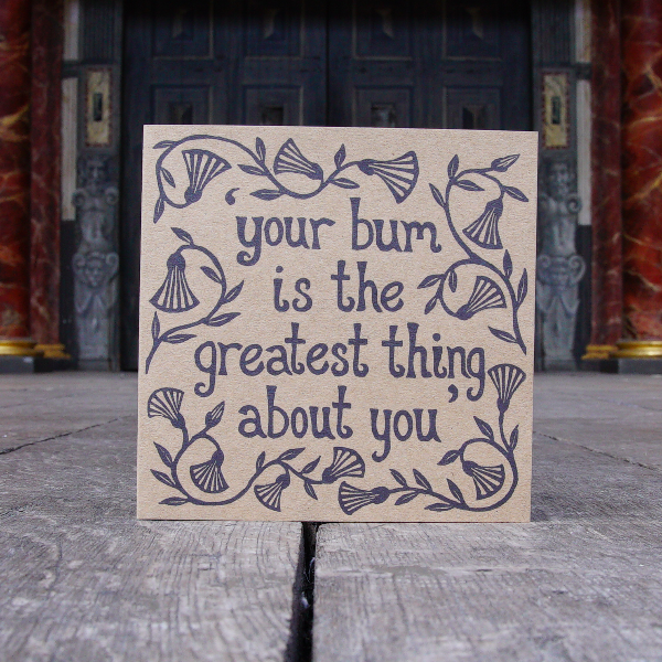 Greetings card made of kraft card with a lino-print in black of a quote from Shakespeare play, Measure for Measure (Your bum is the greatest thing about you). The lettering is hand-drawn and is surrounded by leaves and flowers.