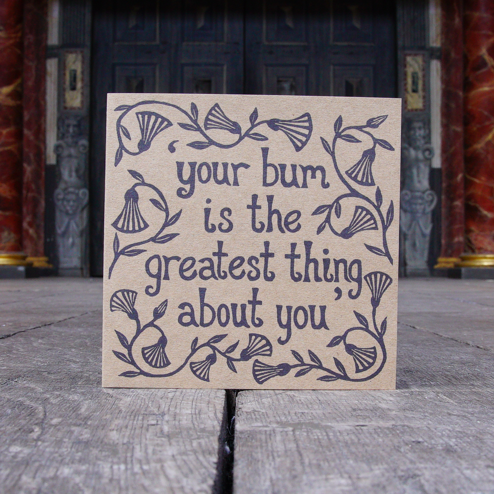Greetings card made of kraft card with a lino-print in black of a quote from Shakespeare play, Measure for Measure (Your bum is the greatest thing about you). The lettering is hand-drawn and is surrounded by leaves and flowers.