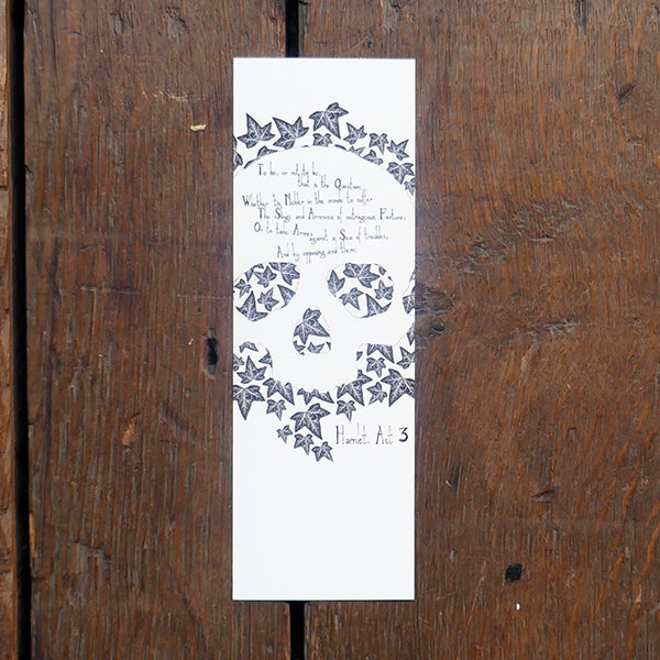 Large card bookmark with a white background, printed with a black and white image of a human skull on a bed of ivy leaves. A quote from Shakespeare play, Hamlet , "To be or not to be..." is printed over the forehead of the skull.