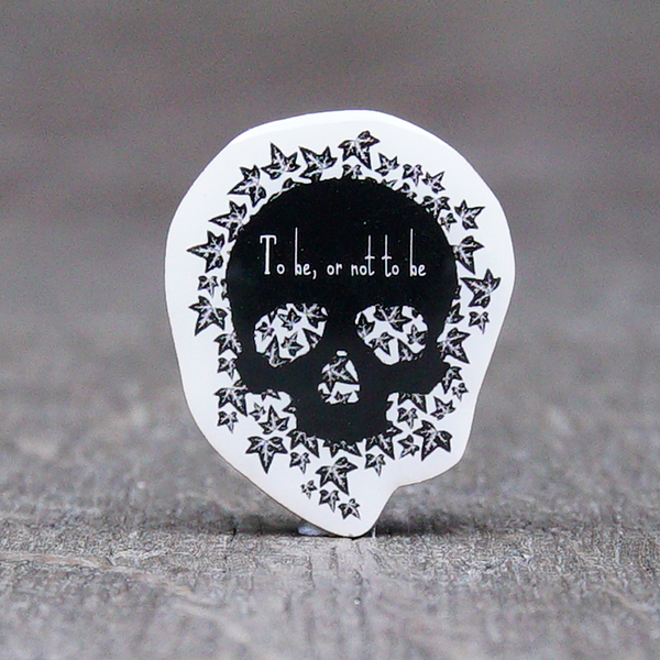 A lapel badge with an image and quote from Shakespeare play, Hamlet. The image consists of a simple black human skull without a bottom jaw and looking straight at the viewer. The skull sites on a bed of ivy leaves, also black but with grey and white details. The background to the badge is white. Across the forehead of the skull is the quote, 
