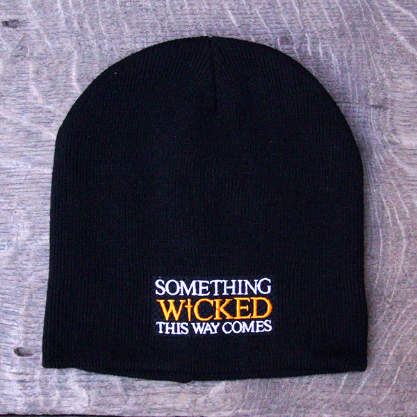 Black beanie hat with an embroidered quote on the front from Shakespeare play, Macbeth, "something wicked this way comes." The quote is in white capital letters with the word 'wicked' in bright orange capital letters.