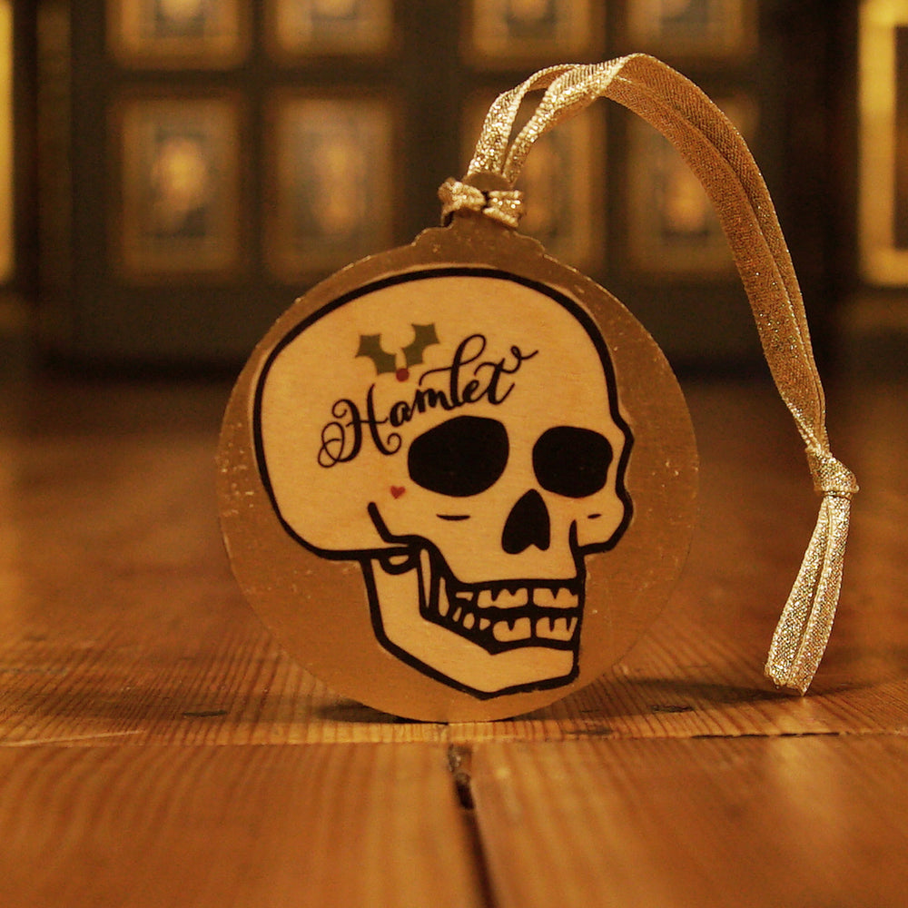 
                  
                    Flat circular wooden Christmas decoration with a printed image of a line-drawn human skull in three-quarter profile. The skull is printed in black and has Hamlet written on its forehead along with a sprig of holly in traditional green and red. A small red heart sits just below the eye socket of the skull. The background is hand-painted in metallic gold. The decoration has a gold ribbon.
                  
                