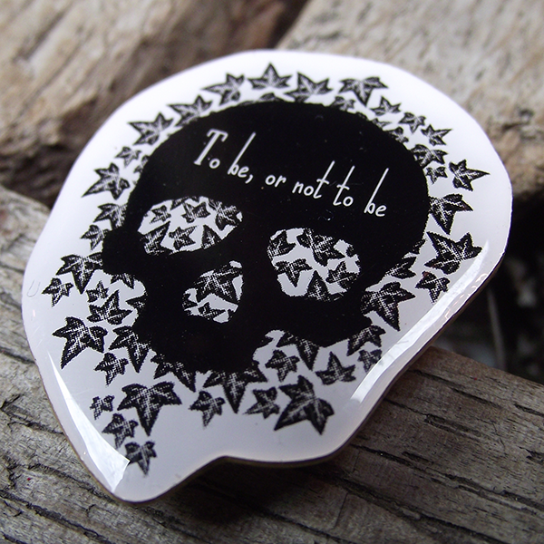 
                  
                    A lapel badge with an image and quote from Shakespeare play, Hamlet. The image consists of a simple black human skull without a bottom jaw and looking straight at the viewer. The skull sites on a bed of ivy leaves, also black but with grey and white details. The background to the badge is white. Across the forehead of the skull is the quote, "to be, or not to be", in spidery, elongated hand-drawn lettering
                  
                