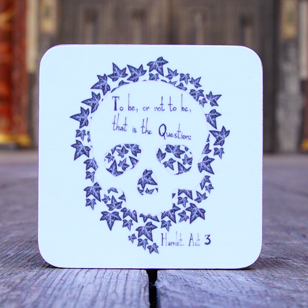 Square melamine coaster with rounded corners. The coaster is white with an image printed in greys and blacks of a simple human skull looking face on. the skull has a background of ivy leaves. Across the forehead of the skull is a quote from Shakespeare play, Hamlet, "To be, or not to be, that is the question?" in hand-drawn lettering.