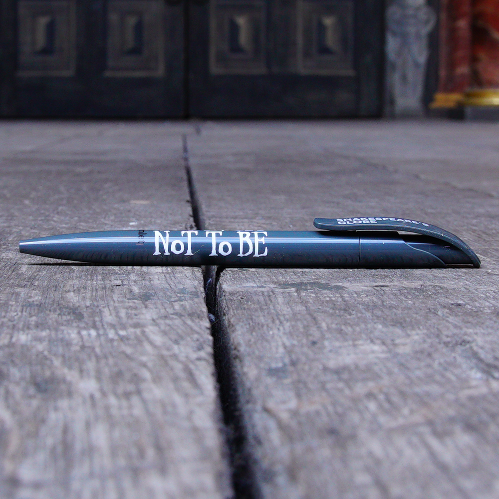 Grey ballpoint pen with a printed quote on the barrel. The quote from Shakespeare play, Hamlet, is printed in white hand-drawn letters (To be or not to be) The name of the play is printed in black next to the quote. The Shakespeare's Globe logo is printed in white on the clip.