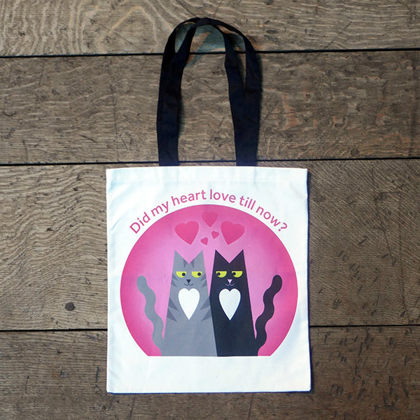White cotton bag with black mid-length handles. A large print is on both sides of two cartoon cats, one a grey tabby , the other black. Both cats are smiling and looking lovingly at each other, both have a heart shaped white chest patch. They sit together in front of a pink circle with love hearts above them. Around the circle is a quote from Shakespeare play, Romeo & Juliet, 