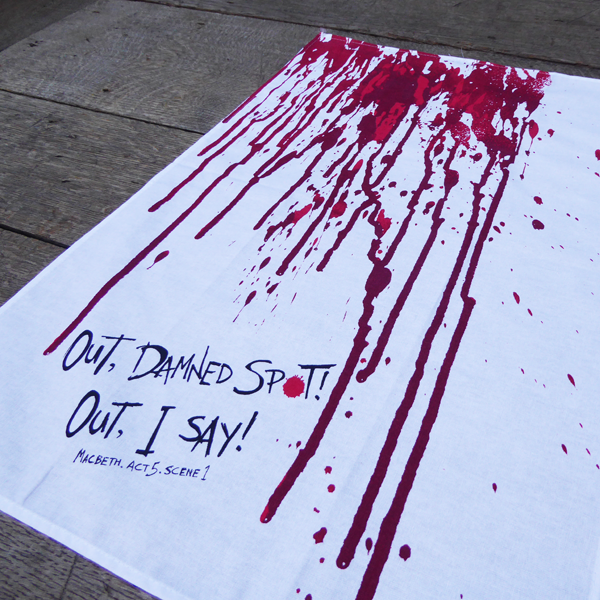 White cotton tea towel with a print of blood dripping down the length of the towel, in reds. At the bottom of the towel is a quote from Shakespeare play, Macbeth (Out. damned spot! Out, I say!) The quote is written in an angry hand-drawn style in black.