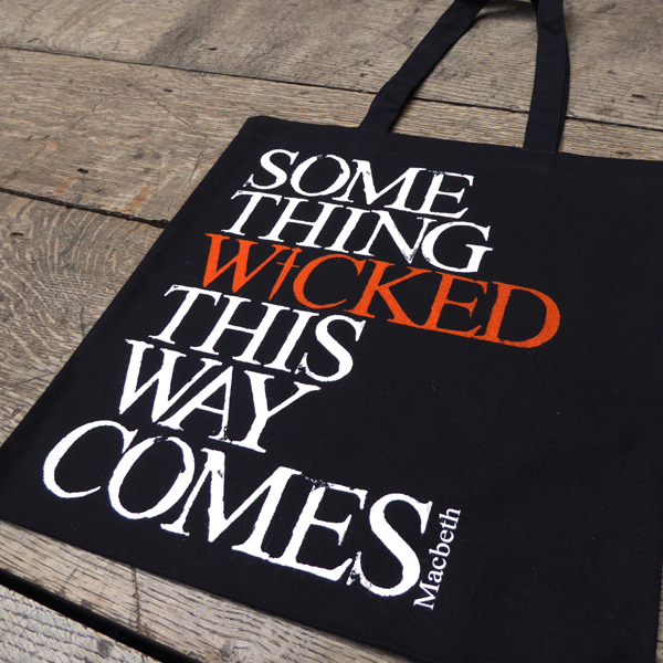 Black cotton bag with mid-length black handles printed with a quote from Shakespeare play, Macbeth (Something wicked this way comes). The lettering is bold serifed capital letters which are slightly distressed printed in white, except for 'WICKED) which is printed in orange. The 'I' of wicked is replaced by a dagger.
