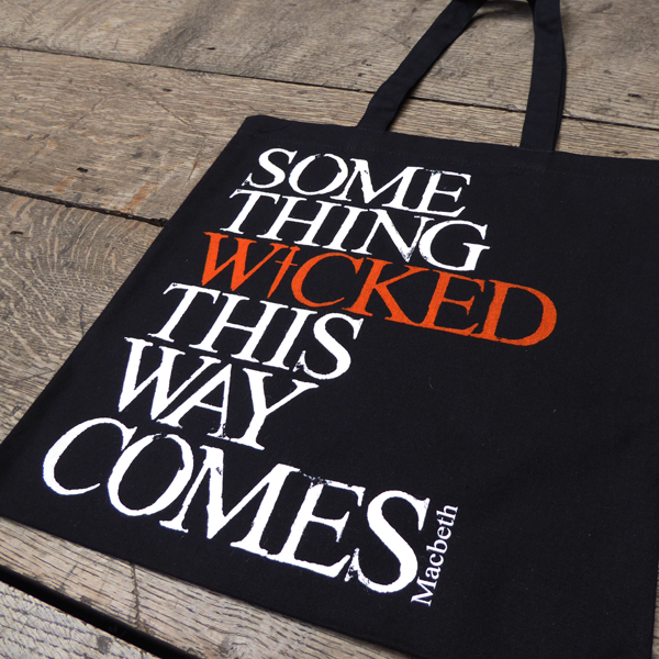 
                  
                    Black cotton bag with mid-length black handles printed with a quote from Shakespeare play, Macbeth (Something wicked this way comes). The lettering is bold serifed capital letters which are slightly distressed printed in white, except for 'WICKED) which is printed in orange. The 'I' of wicked is replaced by a dagger.
                  
                