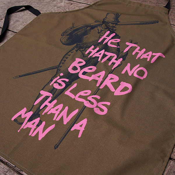Olive green cotton apron with black neck loop and ties. The apron is printed with an etching in black of a man in Stuart dress and a fine beard. Over this is printed a quote from Shakespeare play, Much Ado About Nothing, "he that hath no beard is less than a man" in dusty pink hand-drawn letters