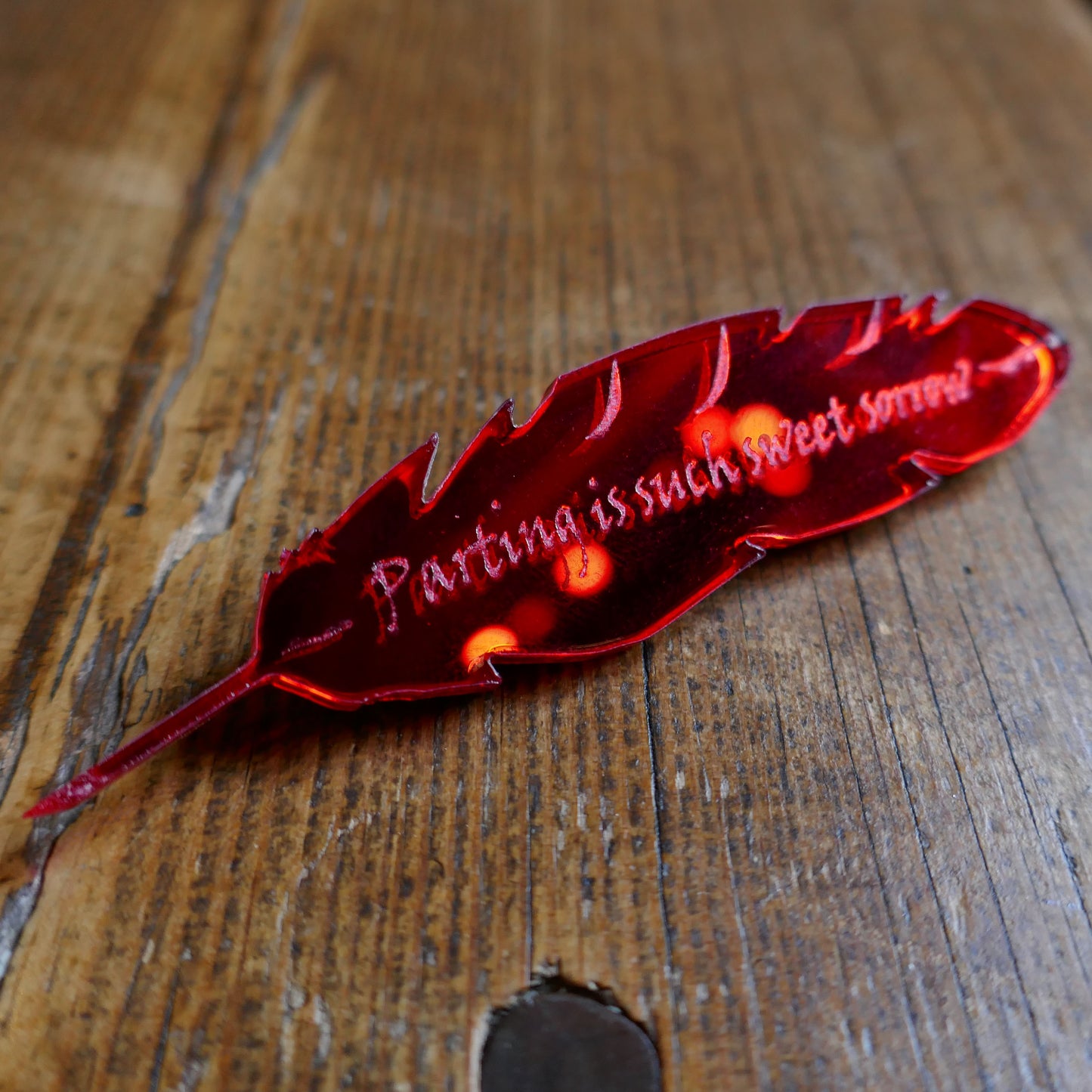 
                  
                    Red mirrored acrylic brooch shaped like a quill and engraved with a quote from Romeo and Juliet
                  
                