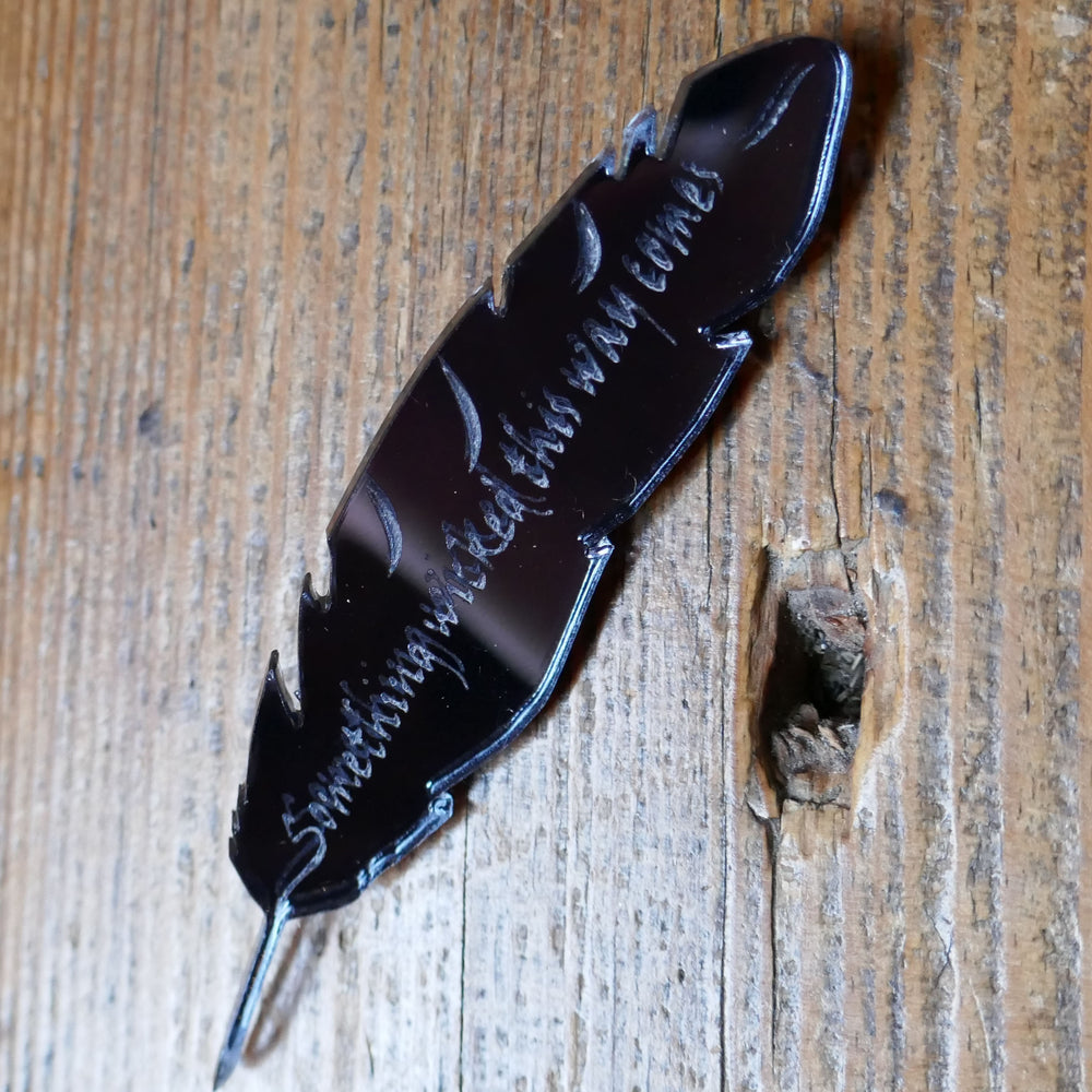 
                      
                        Black, mirrored acrylic brooch in the shape of a quill, engraved with a quote from Shakespeare play, Macbeth, "something wicked this way comes".
                      
                    