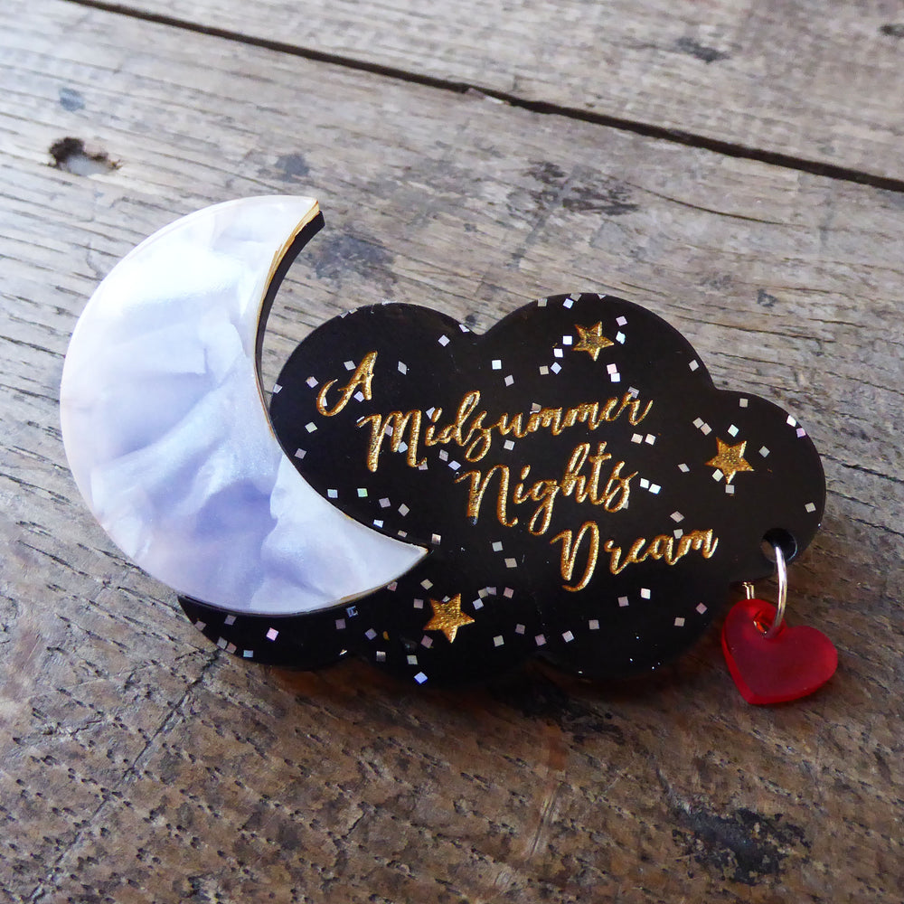 Brooch made of coloured acrylic Perspex. A black cloud with silver glitter forms the background and is engraved with the title of Shakespeare play, A Midsummer Night's Dream in gold, cursive lettering. The cloud is pierced on one side with a small metal ring. On the ring hangs a transparent red heart. On the other side of the cloud is a Perspex cresent moon which looks as though it is made of mother-of-pearl.