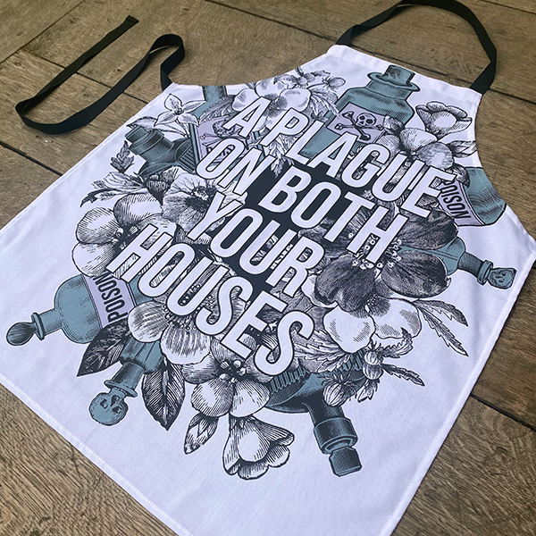 White cotton apron with black neck loop and ties, printed with an image celebrating Shakespeare play, Romeo & Juliet. A background of black and white flowers and green poison bottles with a quote fro the play, "a plague on both you house" printed in white capital letters.
