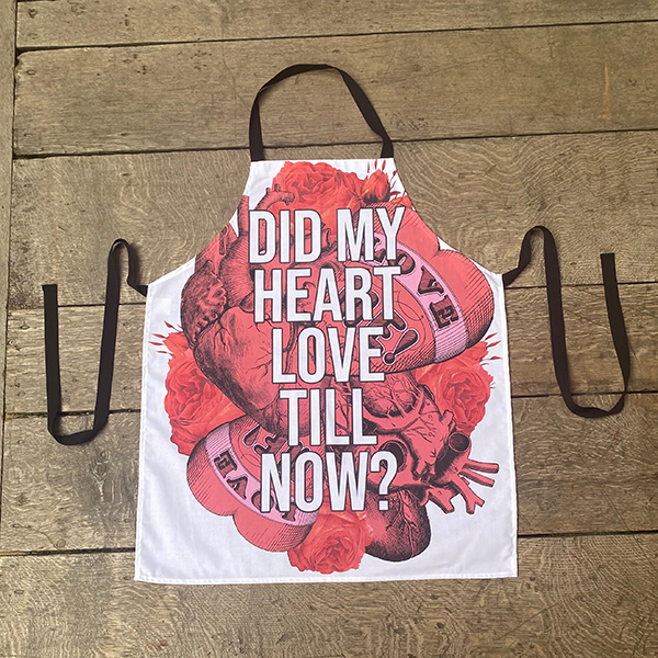 White cotton apron with black cotton neck loop and ties, printed with a image celebrating Shakespeare play, Romeo and Juliet. A background of bright red anatomical and graphic hearts and roses. The graphic hearts have a banner with the words 'love me!' on them. Over the top of this image is a quote from the play 