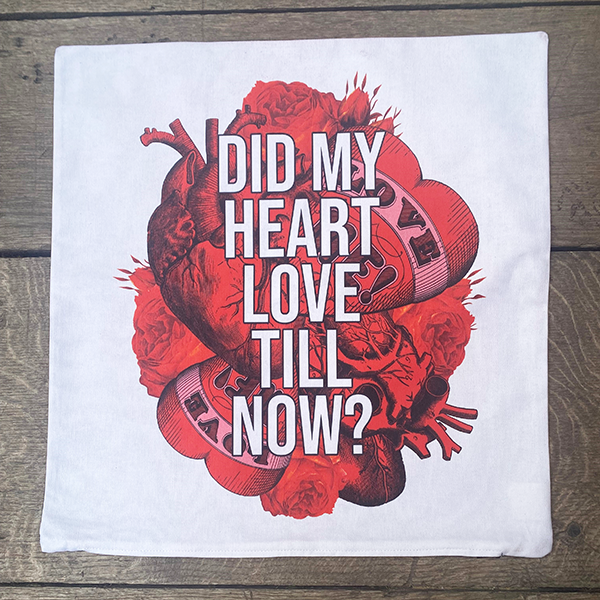 Square white cotton cushion cover, printed with a image celebrating Shakespeare play, Romeo and Juliet. A background of bright red anatomical and graphic hearts and roses. The graphic hearts have a banner with the words 'love me!' on them. Over the top of this image is a quote from the play 