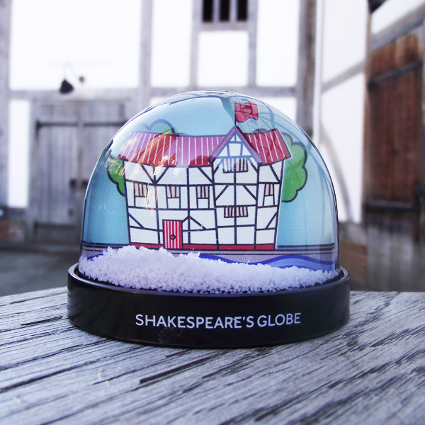 A snow globe containing an illustration of the Globe Theatre. The theatre is drawn in a naive cartoon style with a red thatched red and red door. The theatre walls are cream and the wooden frame is black, yellow light can be seen through the windows. The river flows past the theatre and behind it is a separate panel with green trees on it.