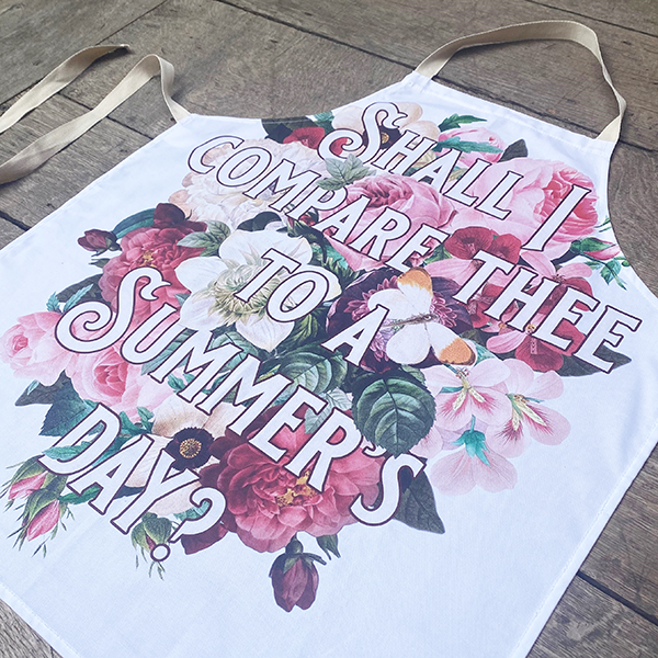 White cotton apron printed with a large print of colourful summer flowers. Ove rthe flowers in a fancy font is the first line from Shakespeare's Sonnet 18 (Shall I compare thee to a summer's day?)
