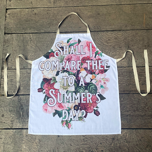 White cotton apron printed with a large print of colourful summer flowers. Ove rthe flowers in a fancy font is the first line from Shakespeare's Sonnet 18 (Shall I compare thee to a summer's day?)