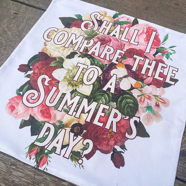 Square white cotton cushion cover printed with a background of colourful summer flowers. Ove rthe top of the flowers is printed the first line of Shakespeare's Sonnet 18 (Shall I compare thee to a summer's day?) in a fancy font.