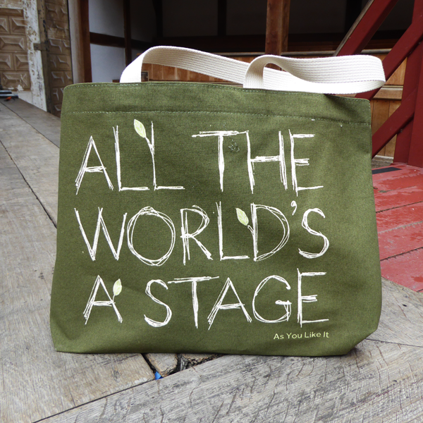 Sturdy sage green tote bag with a quote from Shakespeare play As You Like It (all the world's a stage) printed in white. The lettering is in a hand-drawn scribbled style to represent woodgrain and several of the letters have little lime green leaves growing out from them. The title of the play is printed in lime green under the quote. The bag has white handles. 