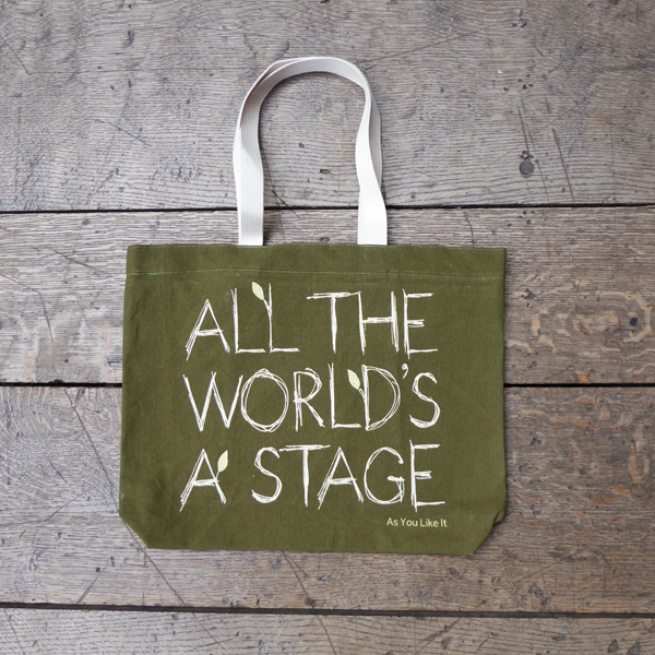 Sturdy sage green tote bag with a quote from Shakespeare play As You Like It (all the world's a stage) printed in white. The lettering is in a hand-drawn scribbled style to represent woodgrain and several of the letters have little lime green leaves growing out from them. The title of the play is printed in lime green under the quote. The bag has white handles. 