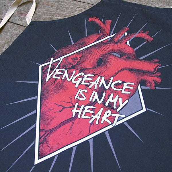 Black cotton apron with natural neck loop and ties, printed with an image celebrating Shakespeare play, Titus Andronicus. A red anatomical heart is partly enclosed by a white diamond frame. Gold sun rays emanate from behind the frame. Across the front of the image is a quote from the play (vengeance is in my heart) in white lettering.