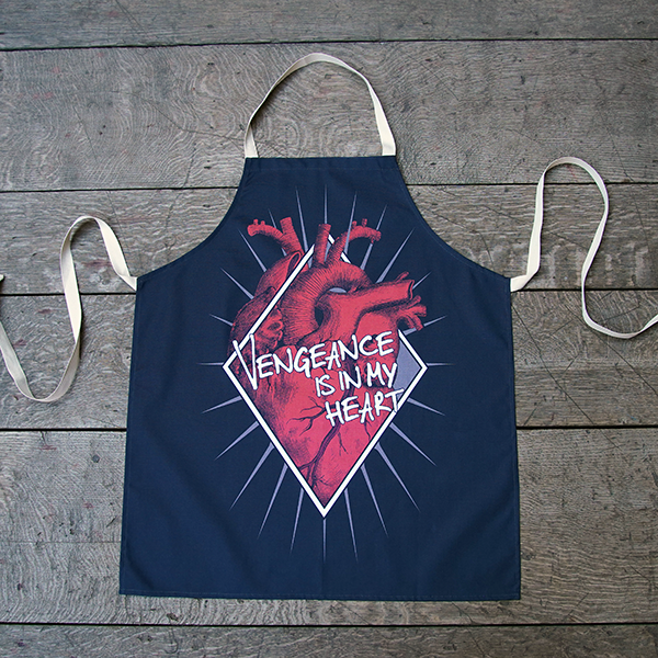 Black cotton apron with natural neck loop and ties, printed with an image celebrating Shakespeare play, Titus Andronicus. A red anatomical heart is partly enclosed by a white diamond frame. Gold sun rays emanate from behind the frame. Across the front of the image is a quote from the play (vengeance is in my heart) in white lettering.