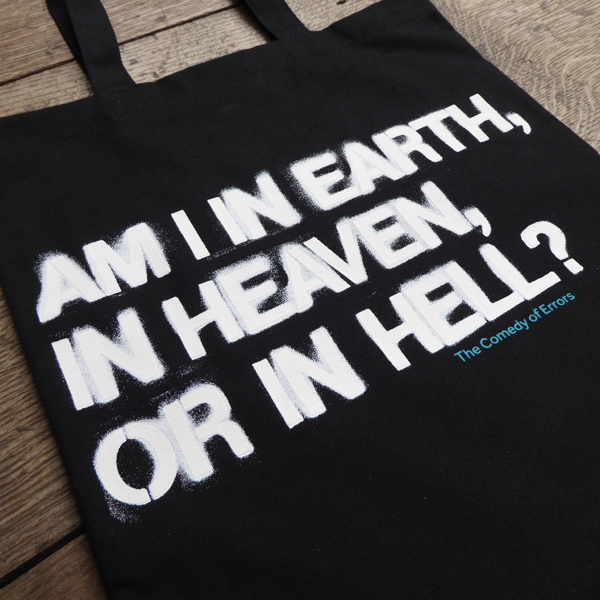 
                  
                    Black cotton bag with black mid-length handles printed with a quote from Shakespeare play, The Comedy of Errors (Am I in earth, in heaven, or in hell?) The letters are printed in bold white san serif capital letters using an expanding dye which gives a sprayed graffiti look. The title of the play is printed in bright blue under the quote.
                  
                