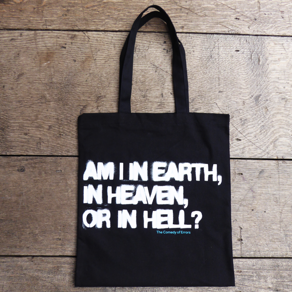 Black cotton bag with black mid-length handles printed with a quote from Shakespeare play, The Comedy of Errors (Am I in earth, in heaven, or in hell?) The letters are printed in bold white san serif capital letters using an expanding dye which gives a sprayed graffiti look. The title of the play is printed in bright blue under the quote.