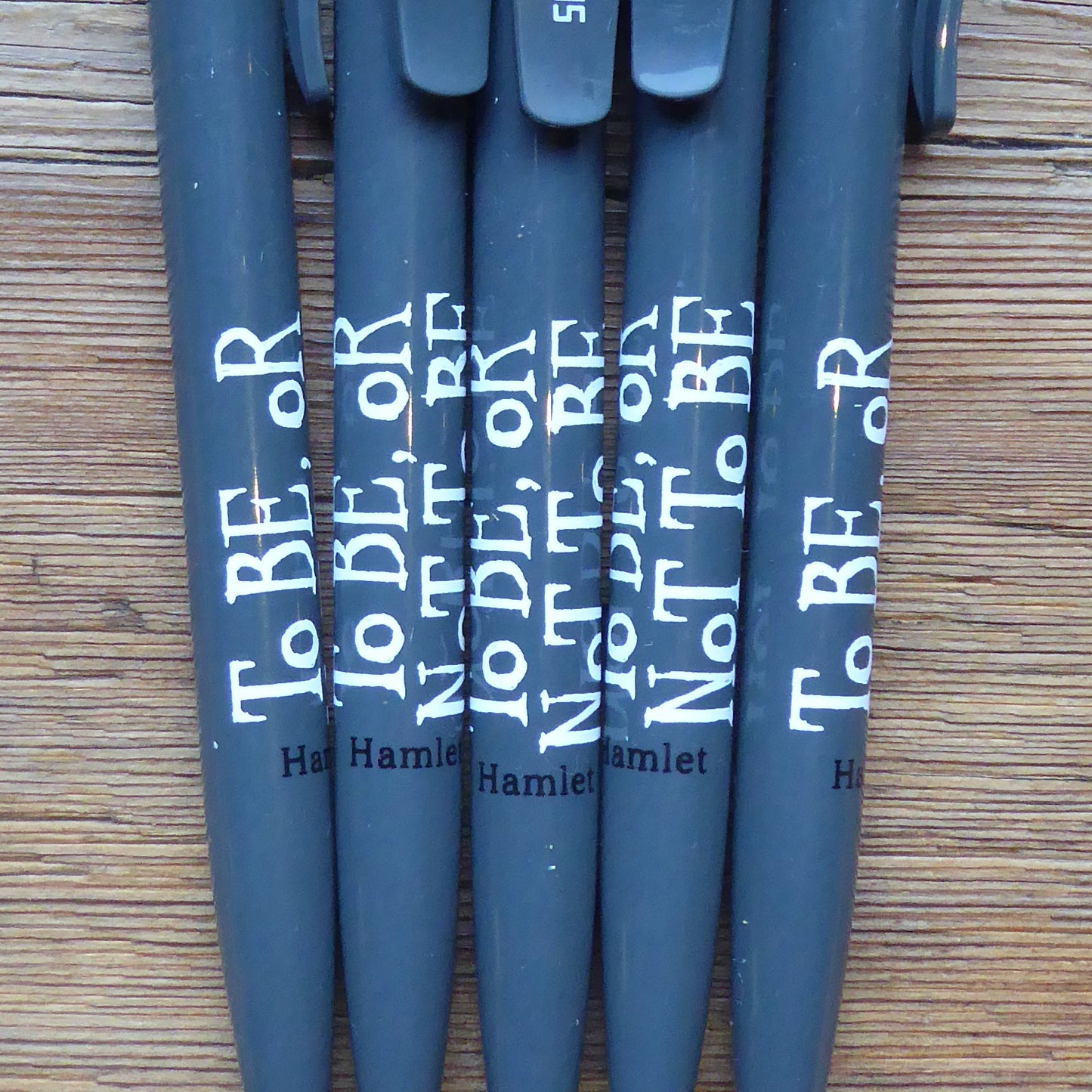 
                  
                    Grey ballpoint pen with a printed quote on the barrel. The quote from Shakespeare play, Hamlet, is printed in white hand-drawn letters (To be or not to be) The name of the play is printed in black next to the quote. The Shakespeare's Globe logo is printed in white on the clip.
                  
                