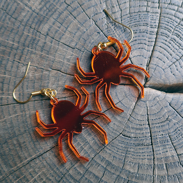 Perspex acrylic drop earring shaped like spiders. The spiders are made from mirror acrylic in orange and have gold coloured metal fixings.